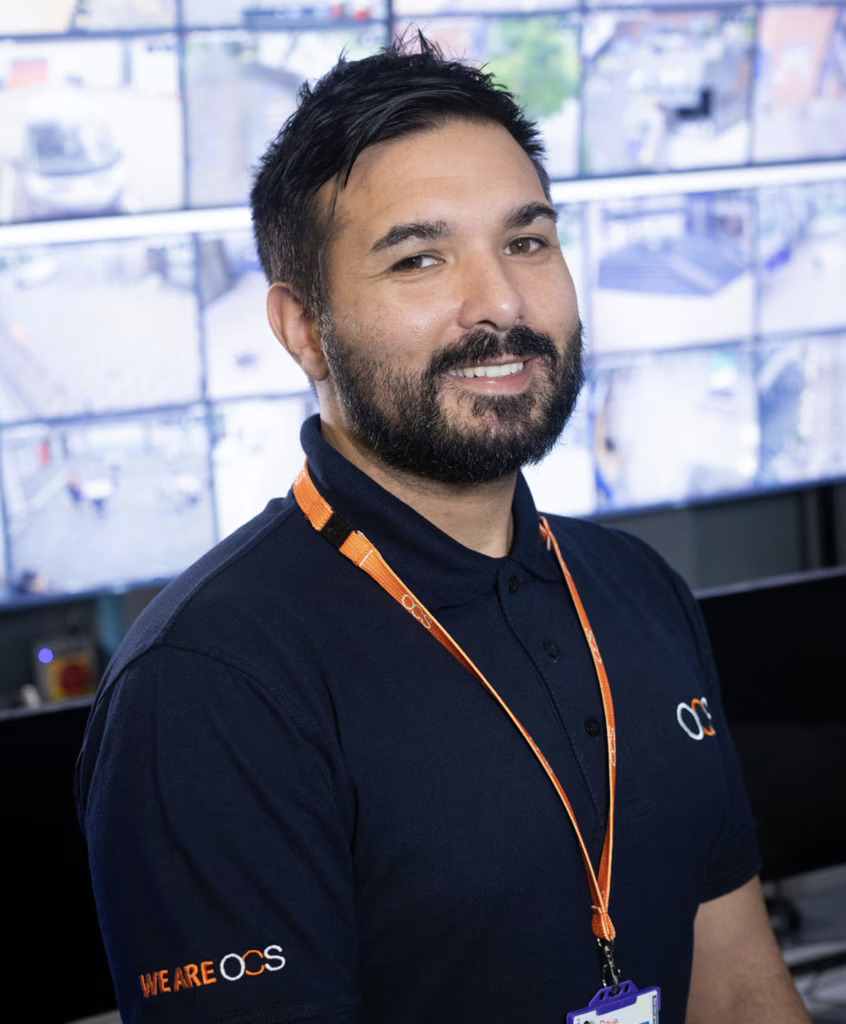 An airport support colleague in a black shirt and orange tie stands in front of a computer screen, ready to assist passengers.