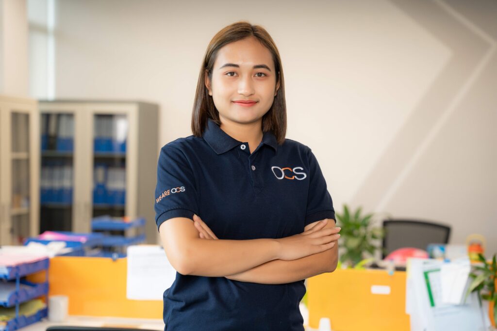 A woman in a blue polo shirt stands confidently in front of a desk, representing her role as a colleague in OCS Cambodia.