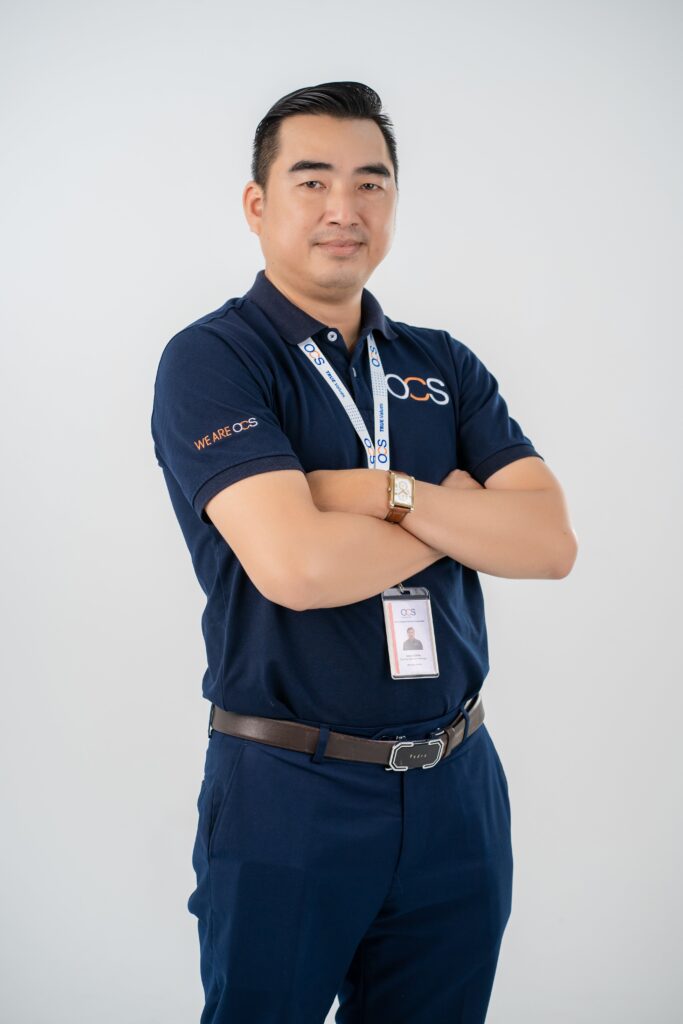 A man wearing a blue polo shirt and blue pants, representing OCS Cambodia, stands confidently in a professional setting.