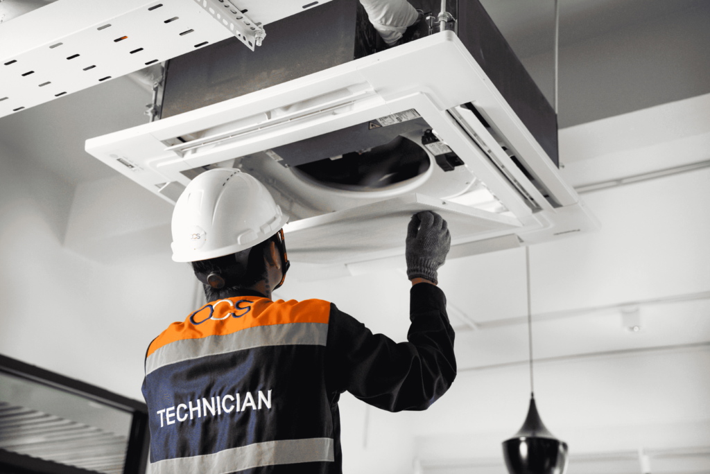 A man in an orange and white hard hat repairs a ceiling fan, representing OCS Cambodia's hard services team.