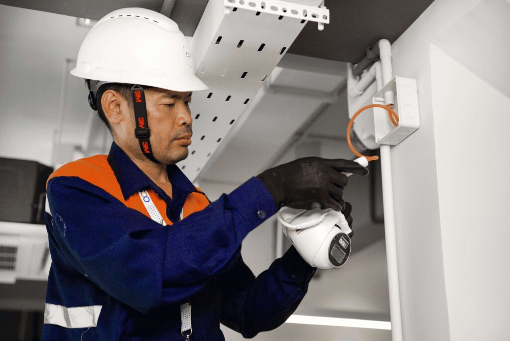 A man in an orange and white hard hat repairs a ceiling fan, representing OCS Cambodia's hard services team.