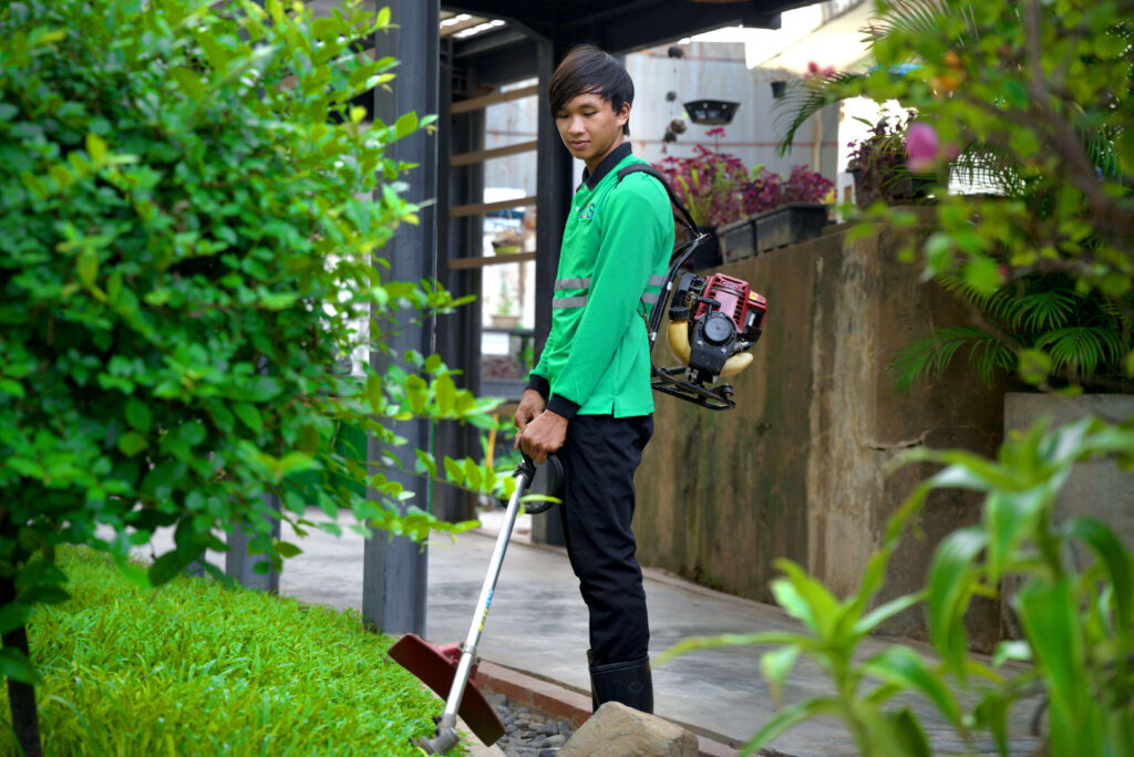 A man in a green shirt and pants uses a garden tool while working on landscaping in Cambodia with colleagues.