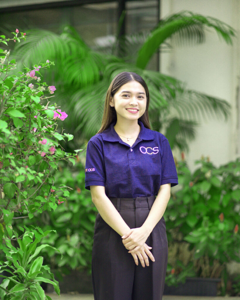 A woman in a purple shirt stands in a landscaped area, representing a colleague from OCS Cambodia.