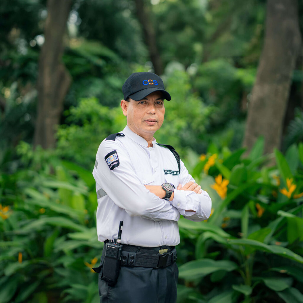 A security officer in uniform stands confidently in front of trees, representing the OCS security team.