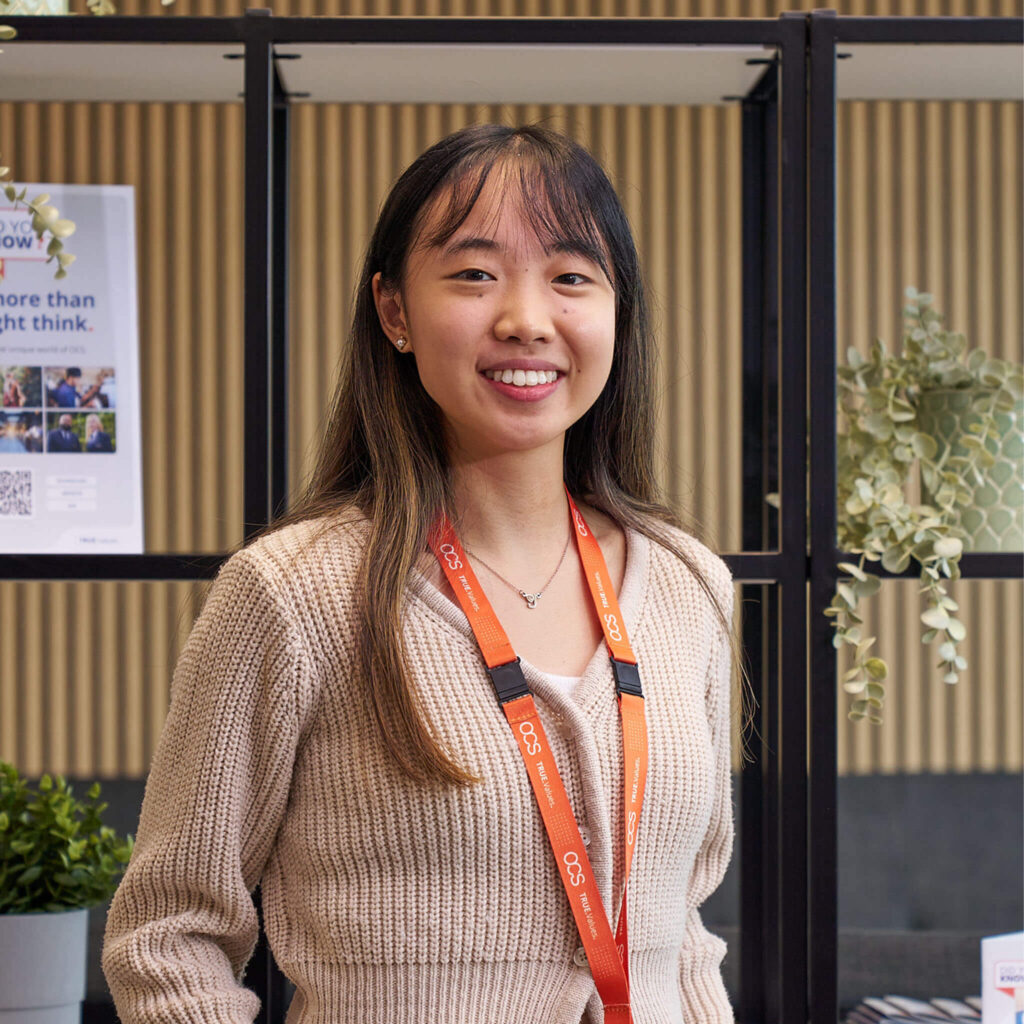 A young woman in a sweater stands confidently in front of a table, representing a new career journey at OCS.