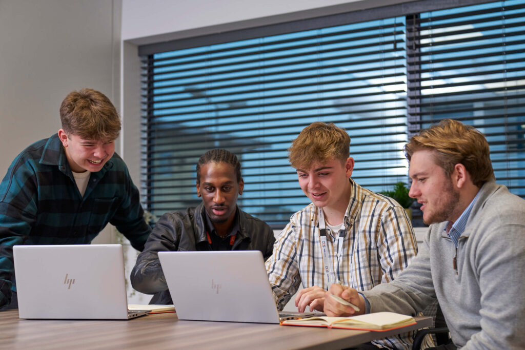 A group of male apprentices engaged in discussion around a table, each using a laptop for collaborative work.