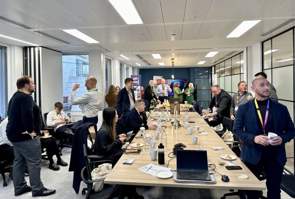 A busy conference room with people standing and sitting around a long table. They are engaged in conversation, enjoying refreshments. The room has large windows and a modern design. Laptops and papers are on the table.