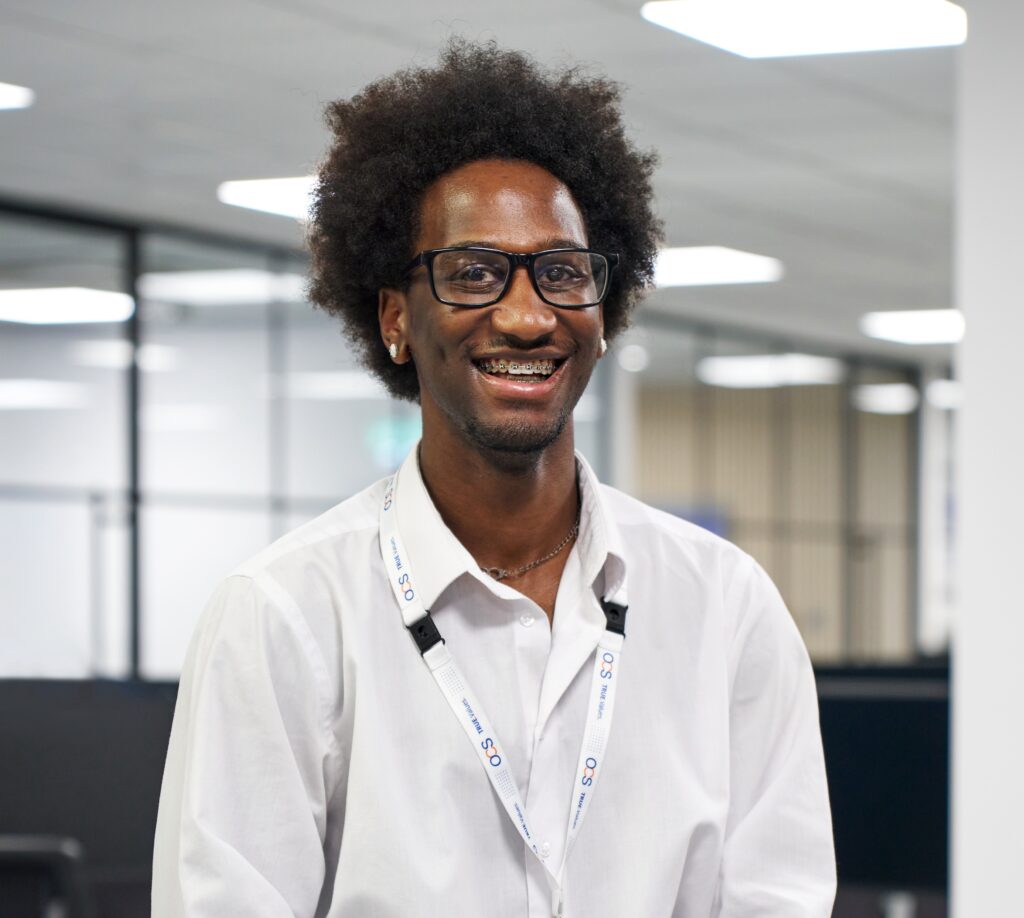 A person with curly hair and glasses is smiling, wearing a white shirt and a lanyard. They are in an office setting with bright overhead lights, black-framed glass partitions, and a blurred background.