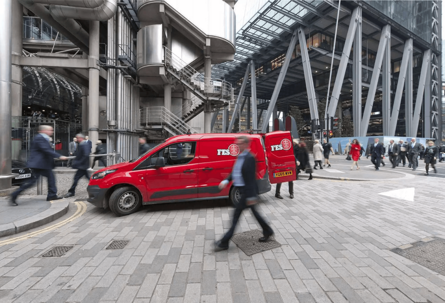 A red FES van is parked on a busy street corner in an urban area with modern buildings. People are walking in various directions, some blurred due to motion. The sky is overcast, enhancing the industrial feel of the scene.