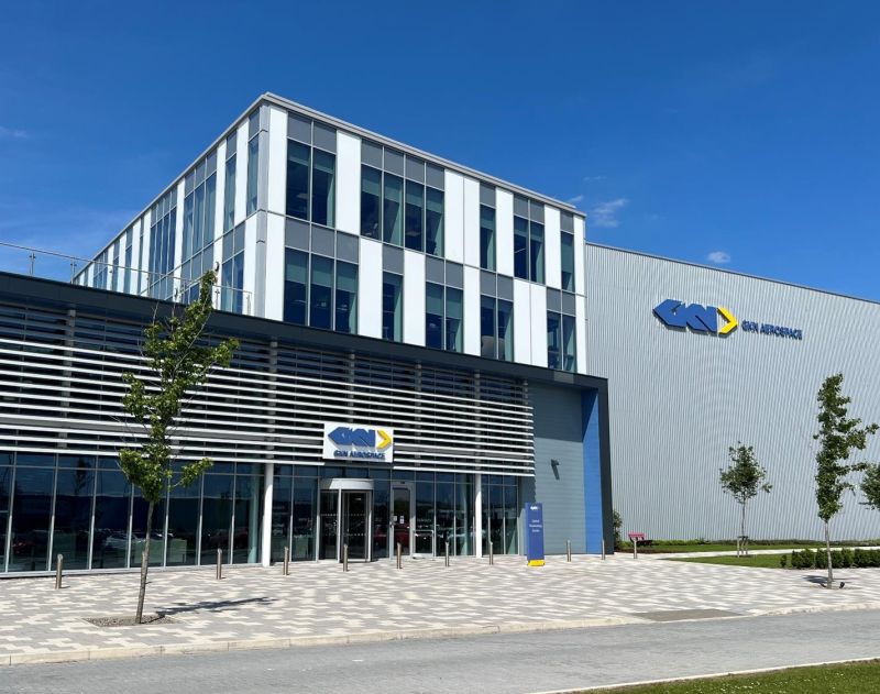 Modern building with glass facade and the logo GEA Aerospace on the exterior. The structure features multiple floors and a sleek design. Trees are planted along the paved walkway under a clear blue sky.