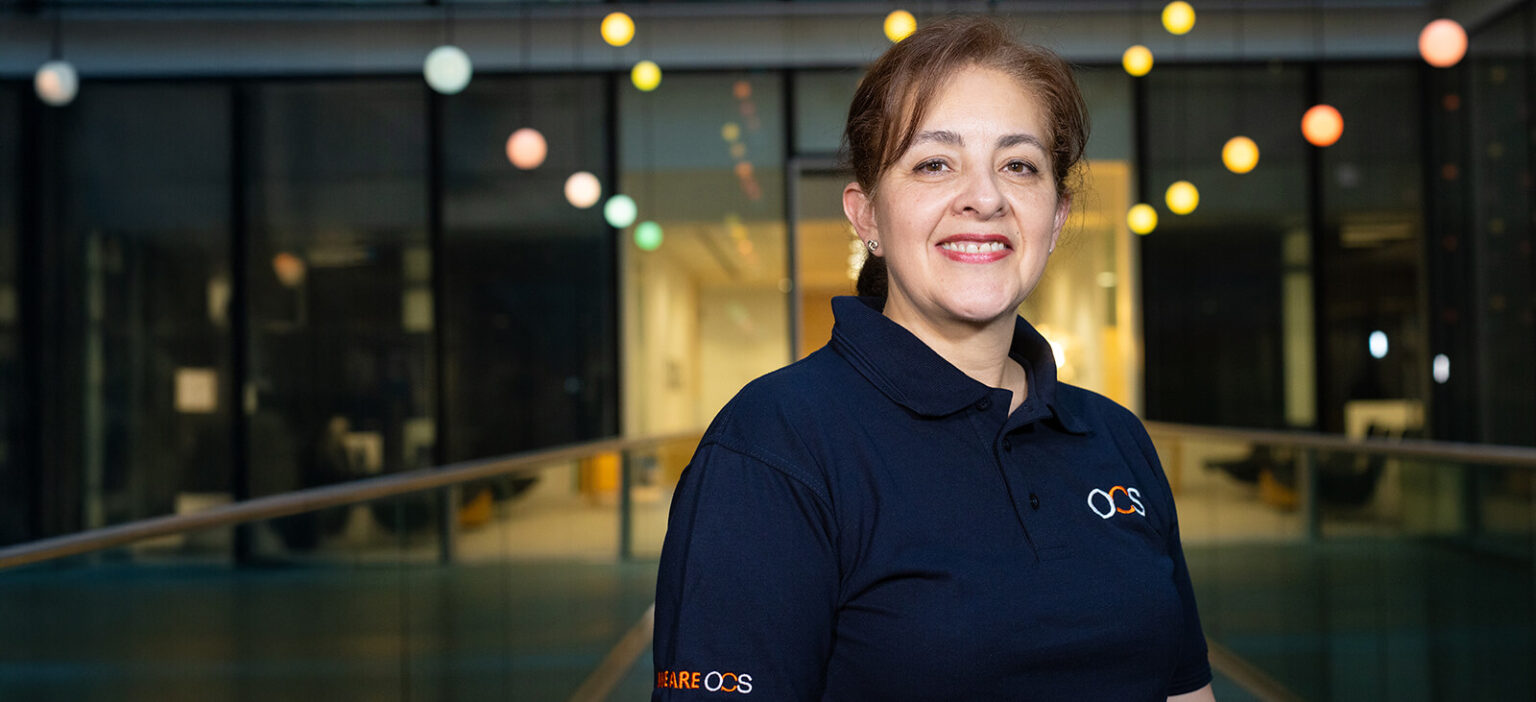 A person with dark hair tied back smiles while standing indoors. They wear a navy blue polo shirt with a logo. Behind them are glass windows and colorful hanging lights.