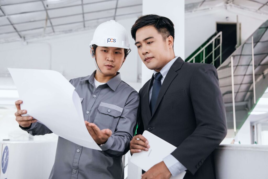 Two men reviewing blueprints in an industrial setting. One, in a hard hat and work attire, points at the papers. The other, dressed in a suit, listens attentively. They appear to be in a discussion about the plans.