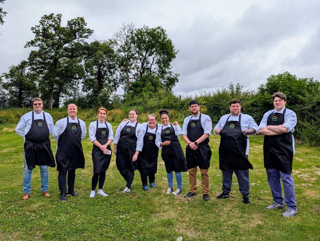 A group of ten people standing on grass, wearing matching aprons. They pose together, smiling, in an outdoor setting with trees and bushes behind them under a cloudy sky.