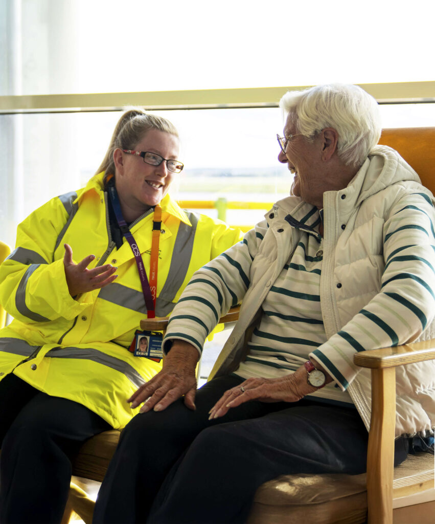 A smiling elderly woman wearing a white vest and a younger OCS PRM colleague in a bright yellow reflective jacket are seated next to each other, engaging in conversation. They are in a sunlit room with a large window in the background.