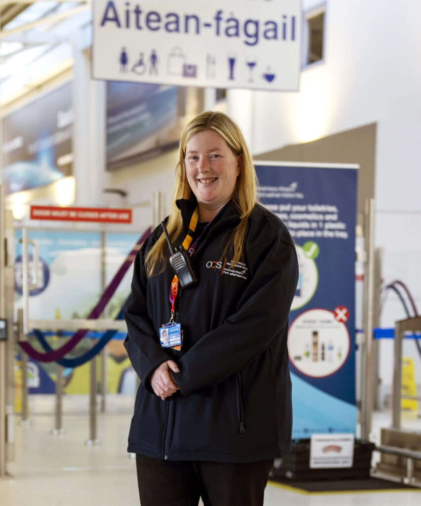 OCS PRM staff member with long hair, wearing a dark jacket and a lanyard, stands smiling in an airport. Signs in Scottish Gaelic and English are visible in the background, along with barriers and informational displays.