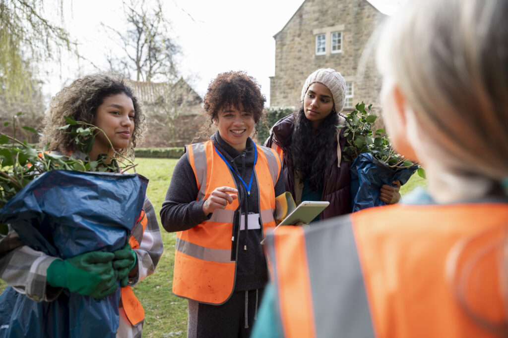 A group of women wearing safety vests and holding gardening tools and bags of leaves gather in a park. One woman is speaking, holding a clipboard. They appear engaged in cleaning or gardening in an outdoor community space.