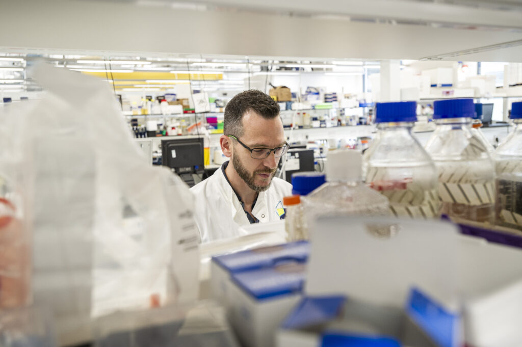 A man in a white lab coat works in a busy laboratory filled with various containers and equipment. He appears focused, surrounded by shelves and counters holding scientific supplies.