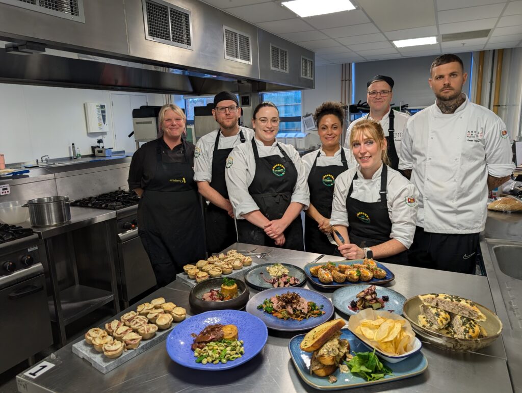 Seven people in a commercial kitchen wearing aprons and chef uniforms stand behind a counter displaying various plated dishes, including meats, vegetables, and pastries. The kitchen features stainless steel appliances and a bright, tiled floor.