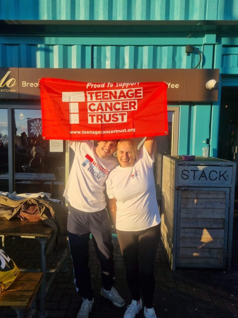 Two people wearing white shirts stand outside, smiling and holding up a red banner that reads Teenage Cancer Trust. The setting appears to be a casual outdoor area with a turquoise building and some benches.