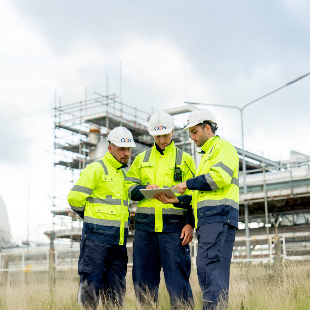 Three construction workers in high-visibility jackets and hard hats stand in a field, reviewing plans on a clipboard. Industrial scaffolding and cloudy skies are in the background.