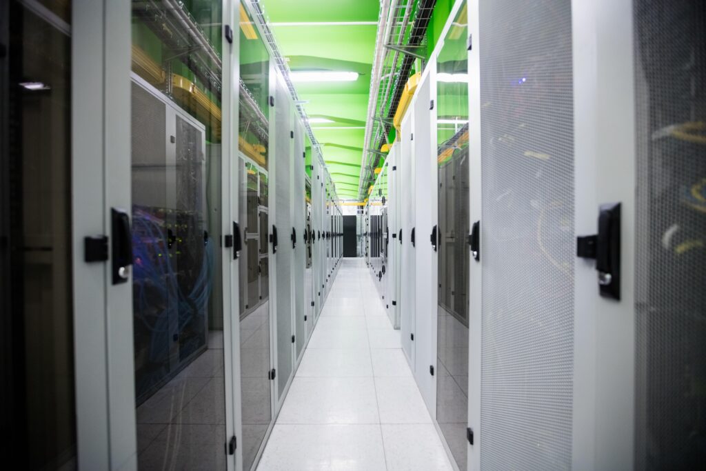 A modern data center hallway lined with tall server racks on both sides. The ceiling is green, and cabling is visible overhead. The floor is white with a glossy finish, reflecting the rows of sleek, metallic cabinets housing electronic equipment.