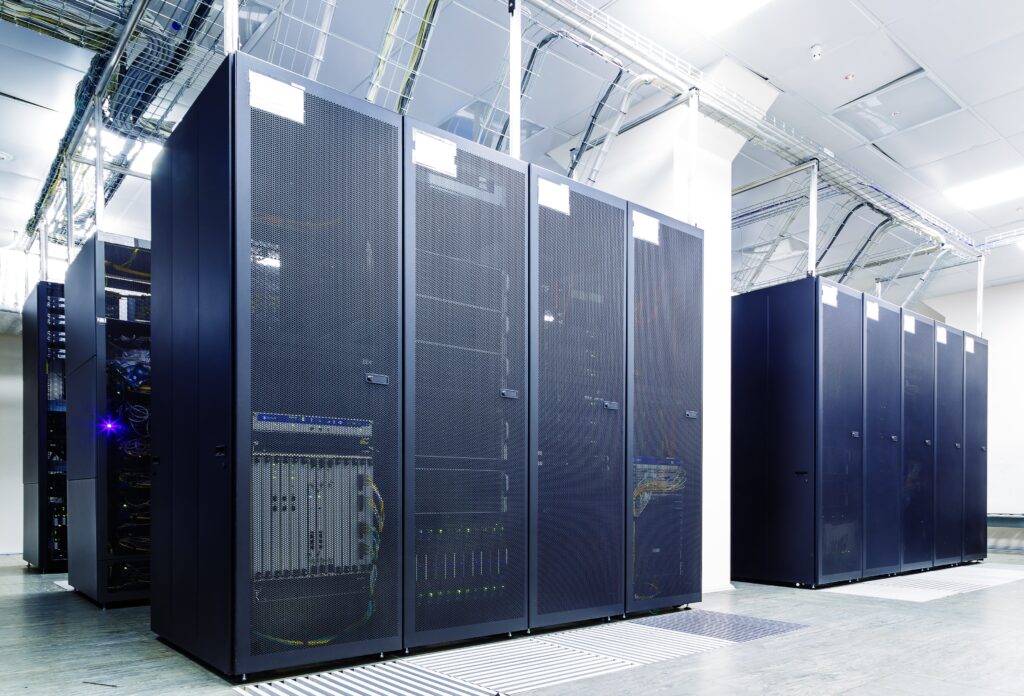 A modern data center with several tall, black server racks lined up in a spacious room. Ceiling cables and bright lighting are visible, reflecting the organized and high-tech environment.