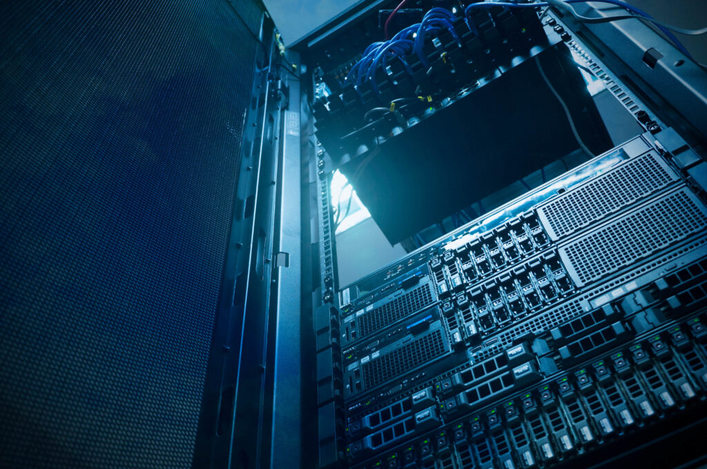 View of an open server rack filled with various electronic components, wires, and servers. The image is taken from a low angle, highlighting the structure and wiring. Cool blue lighting adds a technical atmosphere.