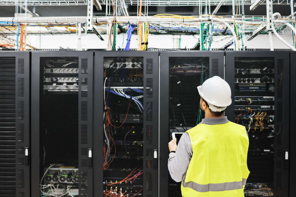 A technician in a high-visibility vest and hard hat inspects server racks filled with cables and equipment in a data center. The focus is on the rear view of the technician as they hold a tablet, surrounded by complex network machinery.