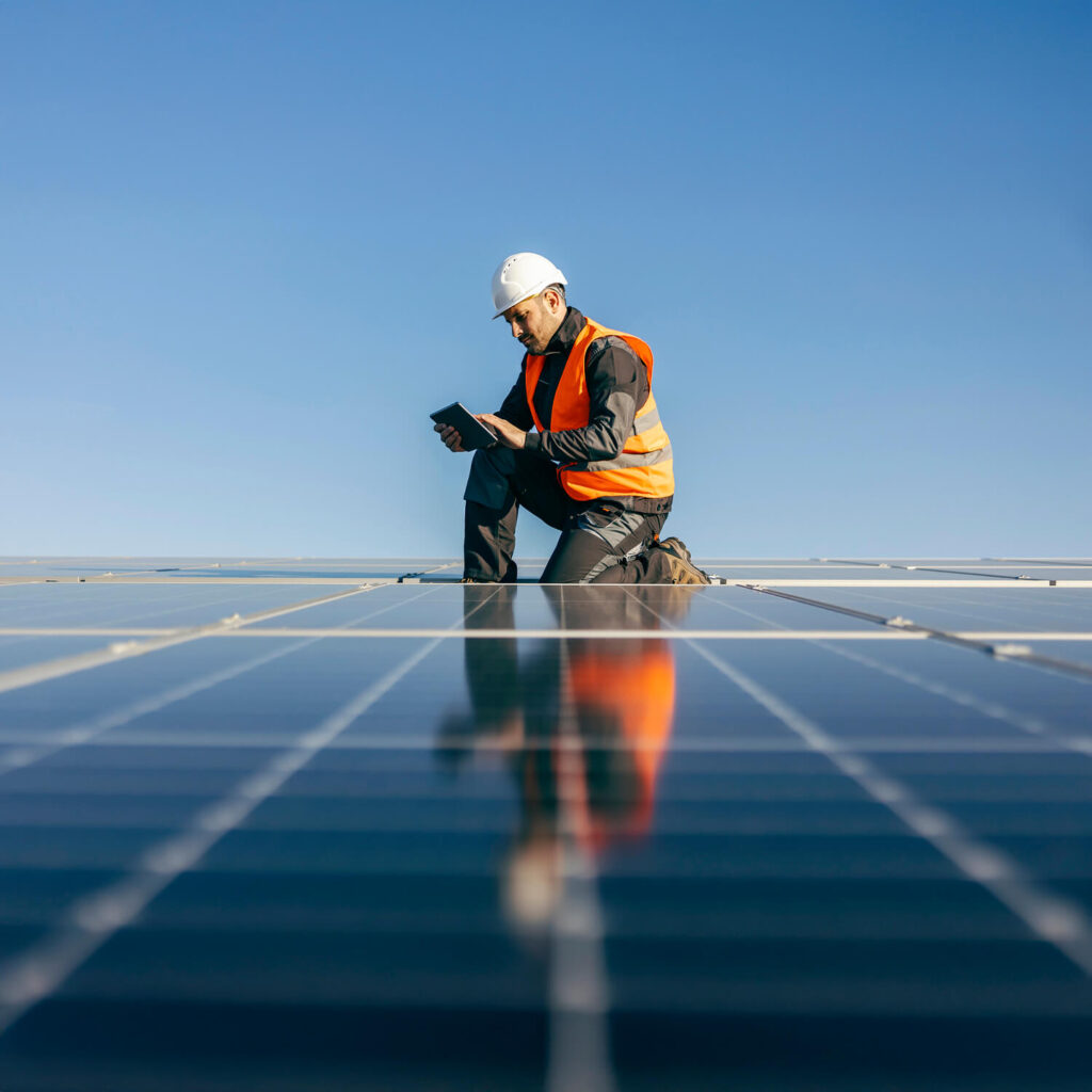 A worker wearing a white hard hat and orange safety vest kneels on a solar panel installation, examining a tablet. The sky is clear and blue, reflecting off the solar panels.