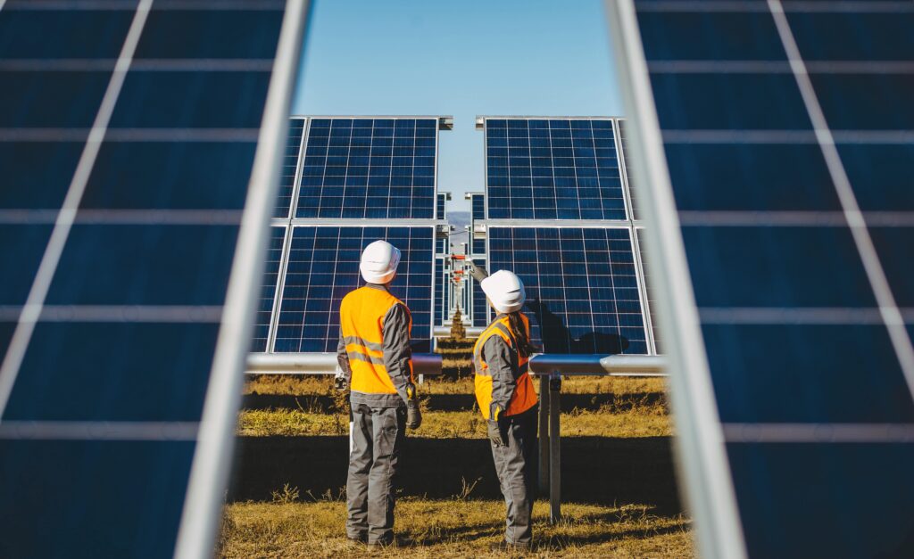 Two workers in orange safety vests and white helmets stand between rows of solar panels, inspecting them. The panels are aligned in a field under a clear blue sky, capturing sunlight for energy.