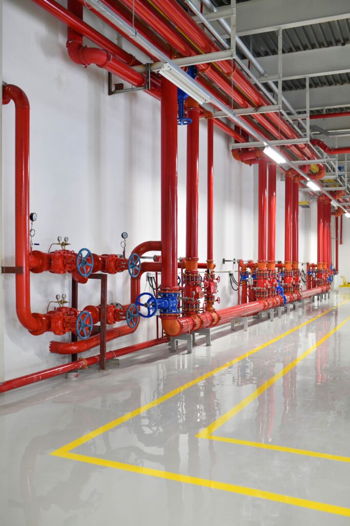 A series of red industrial pipes with blue valves are mounted on a clean white wall inside a facility. The shiny floor reflects the pipes and yellow safety lines are marked on the floor. The ceiling features gray metal beams.