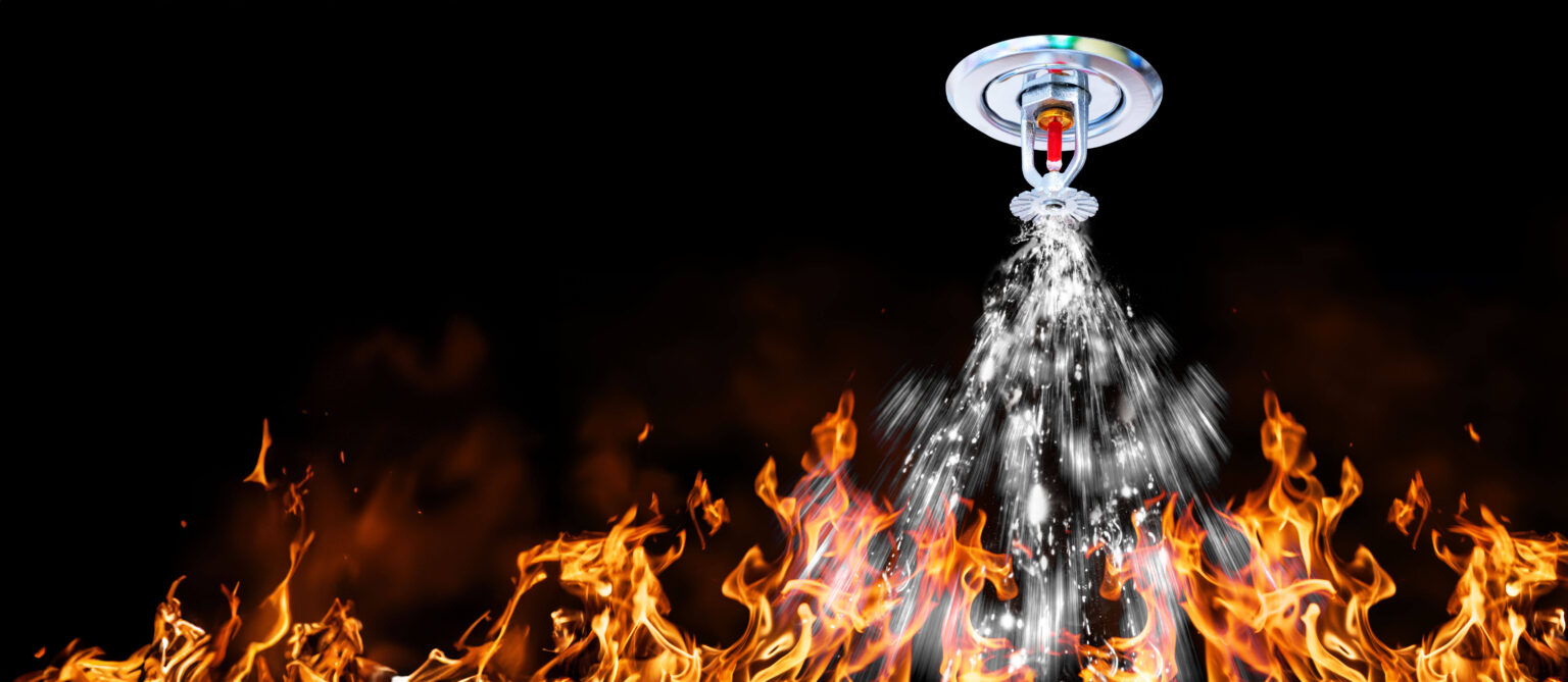 A fire sprinkler activates, spraying water downward. Flames are visible below against a dark background, and the sprinkler emits water in an attempt to extinguish the fire.