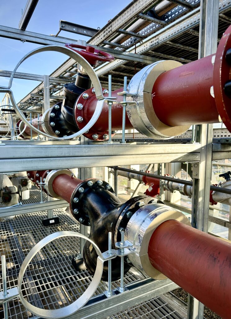 Pipes with red and black protective coatings are secured in a metallic structural frame outdoors. Silver clamps and bolts fasten the pipes, which are positioned parallel to each other, with clear skies visible in the background.