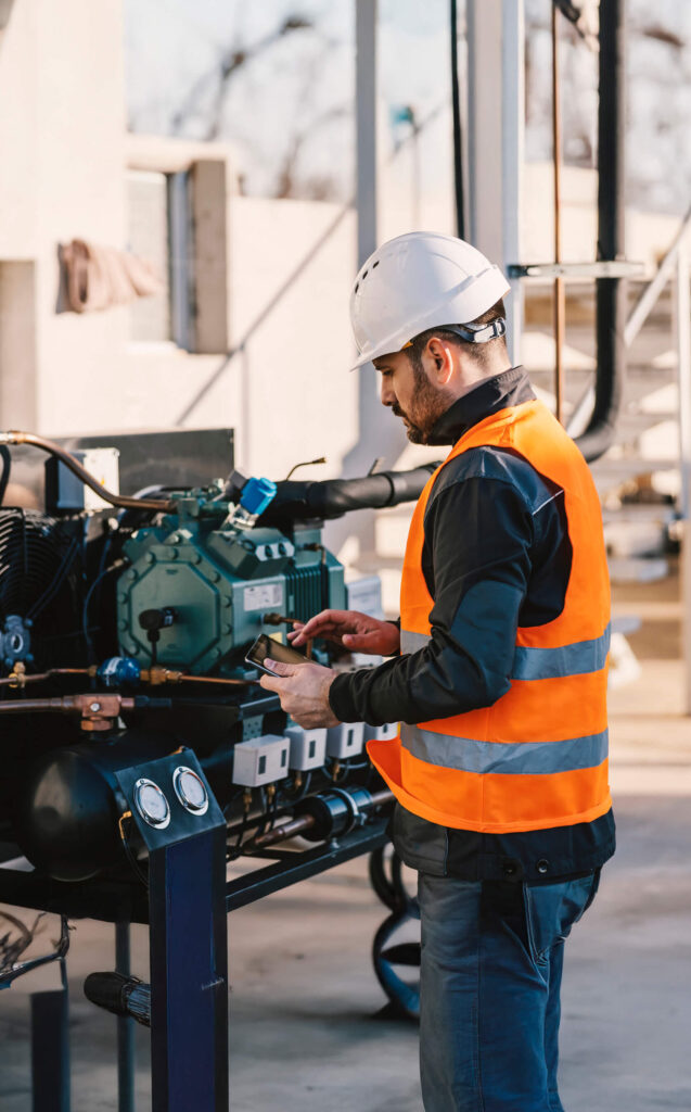 A worker in a white hard hat and orange safety vest is using a smartphone while inspecting machinery outdoors. The equipment includes various pipes and gauges. The setting appears industrial under a clear sky.