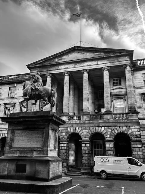 Black and white photo of a historic building with columns, labeled The Signet Library. In front stands an equestrian statue. A van marked OCS is parked nearby. The sky is partly cloudy with a contrail visible.