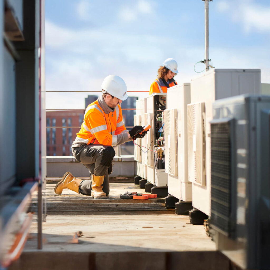 Two individuals wearing hard hats and orange safety vests work on rooftop HVAC units. One person kneels and uses a tool on a unit, while the other stands in the background. A cityscape is visible in the distance. Sky is clear and sunny.