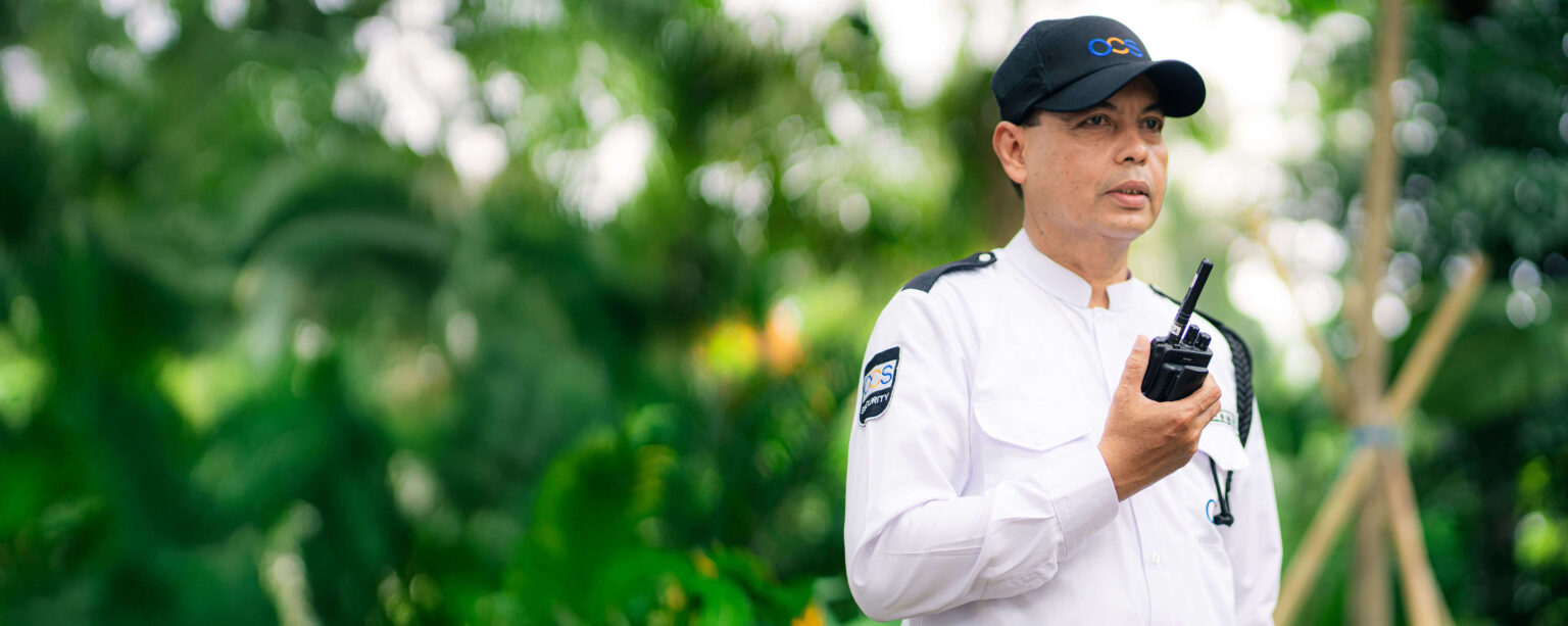 A security guard in a white uniform and black cap holds a walkie-talkie in a lush, green outdoor setting. His uniform has patches, and he appears focused as he looks ahead.