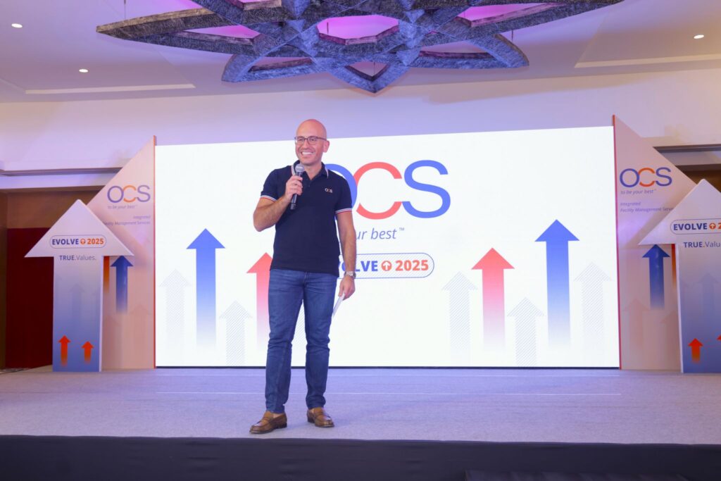 A person in a dark polo shirt and jeans is speaking into a microphone on a stage. Behind them is a screen displaying the text OCS and Evolve @ 2025 with upward-pointing arrows in blue and red.