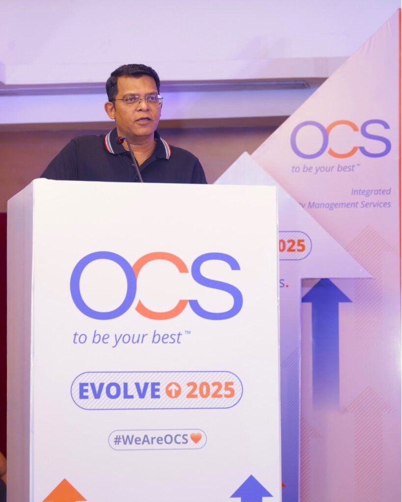 A person stands at a podium with OCS and Evolve 2025 displayed on it. The background features upward arrows, implying progress or growth. The podium also includes the phrase to be your best and #WeAreOCS with a heart symbol.