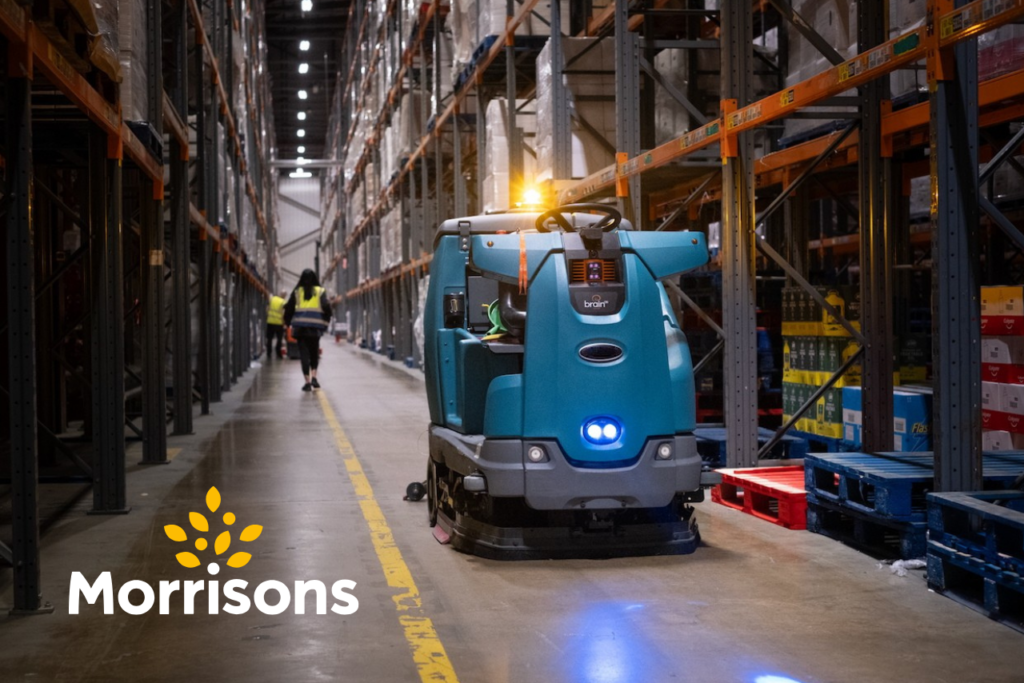 A floor-scrubbing robot operates in a brightly lit warehouse aisle, nearing a worker in the distance. The robot is teal with a glowing light on top. The Morrisons logo is visible in the lower left corner.