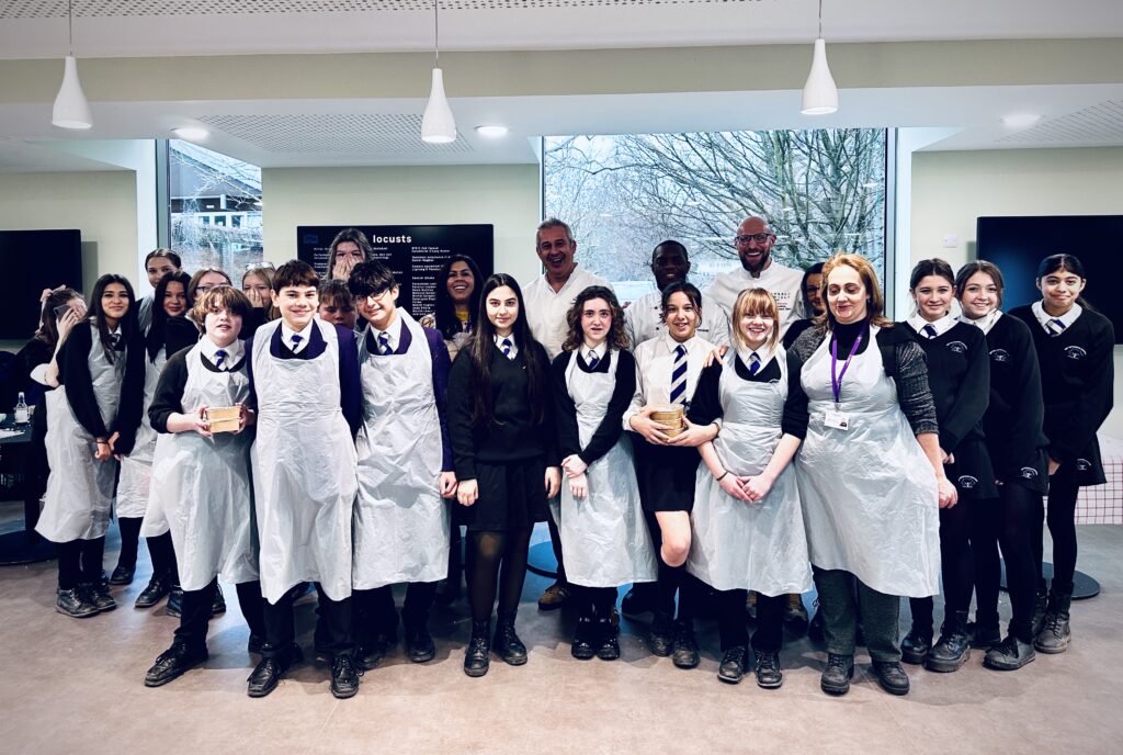 A group of students in uniforms and aprons stand indoors, smiling at the camera. Behind them are adults in chef uniforms. Large windows in the background show trees and a building. The scene appears to be in a cooking or educational setting.