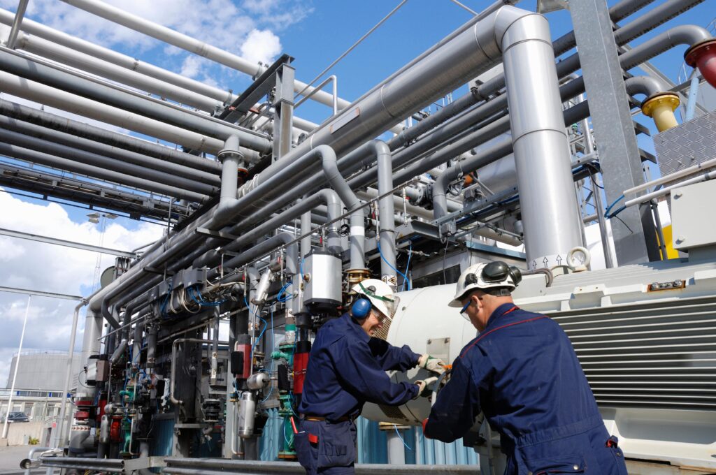 Two workers wearing safety gear inspect machinery in an industrial setting with a network of grey pipes and valves overhead. The sky is partly cloudy in the background.