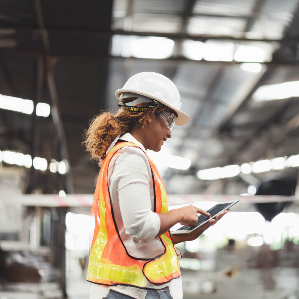 A person wearing a white hard hat, safety goggles, and an orange safety vest is using a tablet in an industrial setting. They are standing indoors with a blurred background of metal beams and machinery.