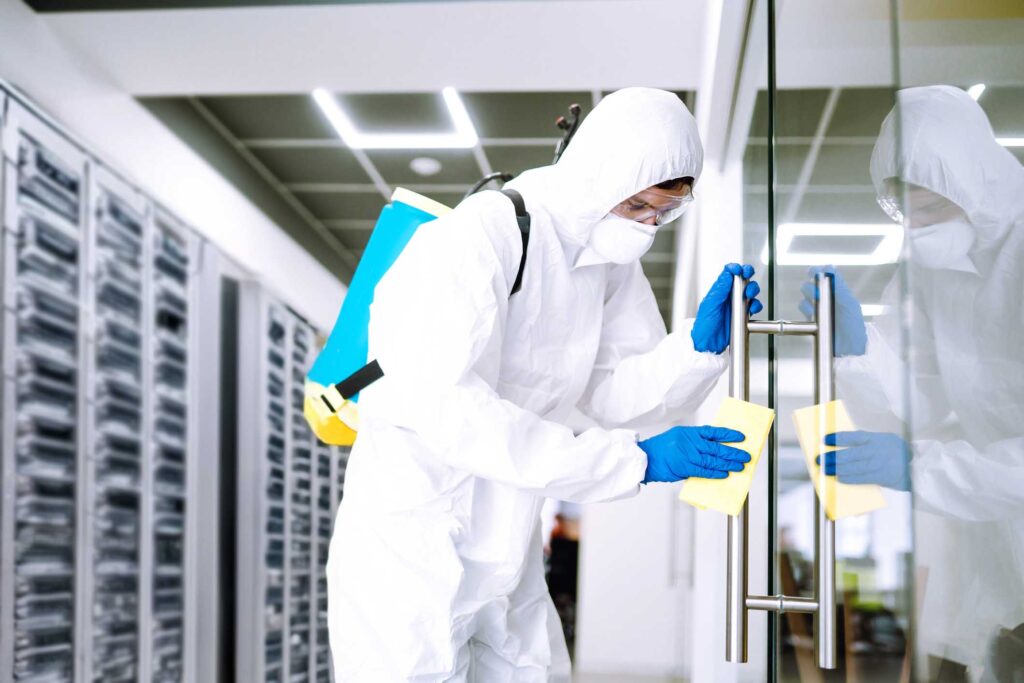 A person in full protective gear, including a white suit, gloves, mask, and goggles, is cleaning a glass door with a yellow cloth. They are in a modern room with a grid-patterned ceiling and silver racks in the background.