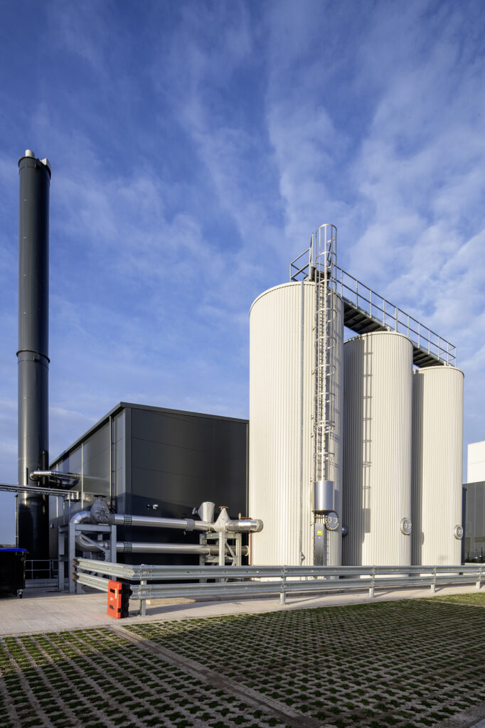 Industrial facility with large cylindrical tanks and a tall chimney against a blue sky with clouds. The area is surrounded by a metal fence and has a paved walkway with grid-pattern grass at the foreground.