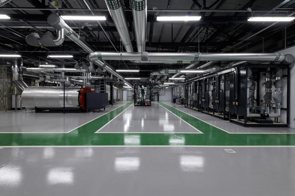 A large industrial facility with a polished floor, featuring metal pipes and ducts on the ceiling. Rows of machinery line the walls, and a large cylindrical tank is on the left. Overhead lights illuminate the space.