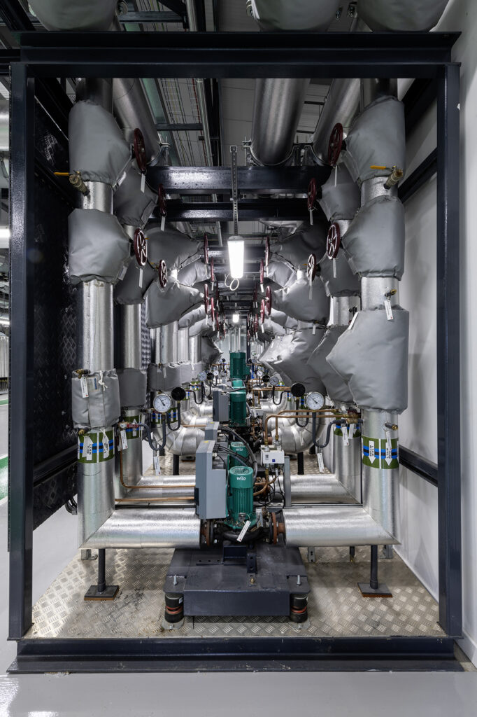 A complex network of insulated industrial pipes and valves is displayed, featuring a central green machine. The pipes are neatly arranged and the equipment is housed within a metal frame, set on a diamond-patterned metal floor.