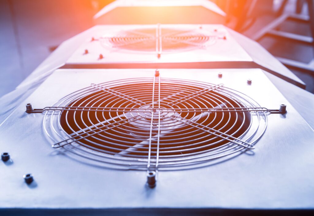 Close-up of industrial cooling fans with protective metal grilles, mounted on rectangular units. The lighting is warm, casting a soft glow on the metallic surfaces. The background is blurred, highlighting the fans as the main focus.