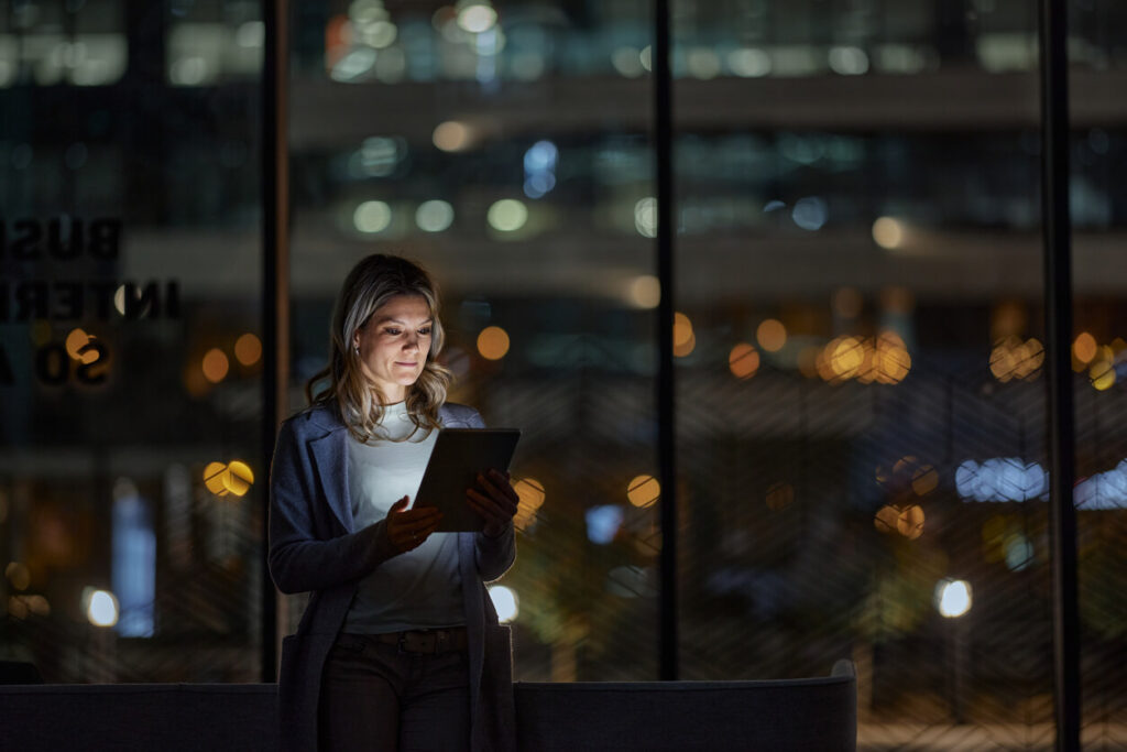 A woman stands indoors using a tablet, illuminated by its light. Shes in front of a large window showcasing a blurred cityscape with glowing lights at night. Her expression appears focused.