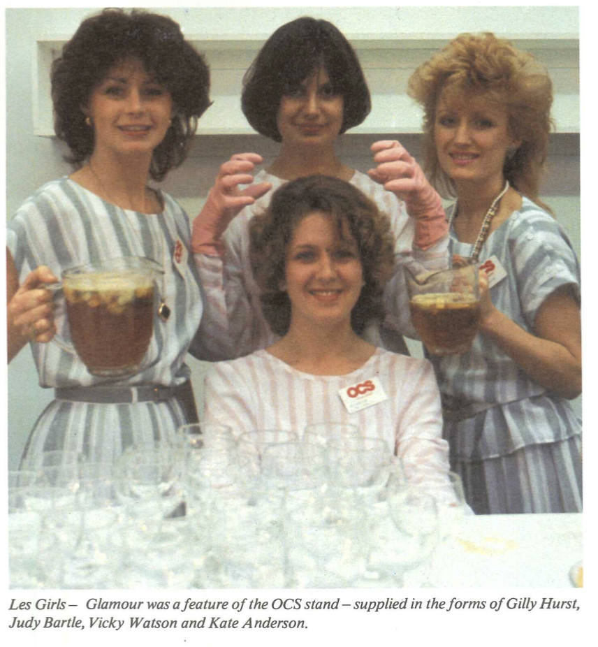 Four women in matching striped outfits stand behind a table filled with empty glasses. Three hold jugs of drinks, while the seated woman holds two. They smile at the camera, with a sign reading OCS on their clothes.